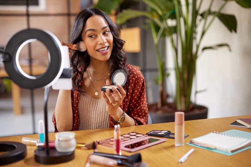 Young woman applying makeup on face with brush while recording vlog on smartphone for social media with LED ring lamp on table. Multiethnic fashion blogger creating cosmetic video. Professional hispanic beauty makeup artist vlogger livestreaming makeup tutorial while reviews a new blush product.